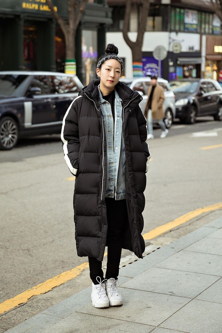Woman layering a long black puffer coat over a denim jacket with white sneakers on the street