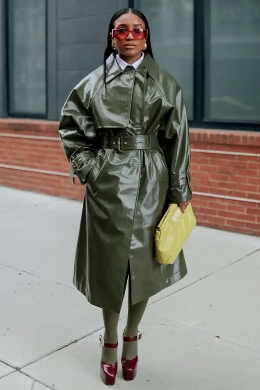 Shiny olive patent trench coat with red heels and a yellow clutch
